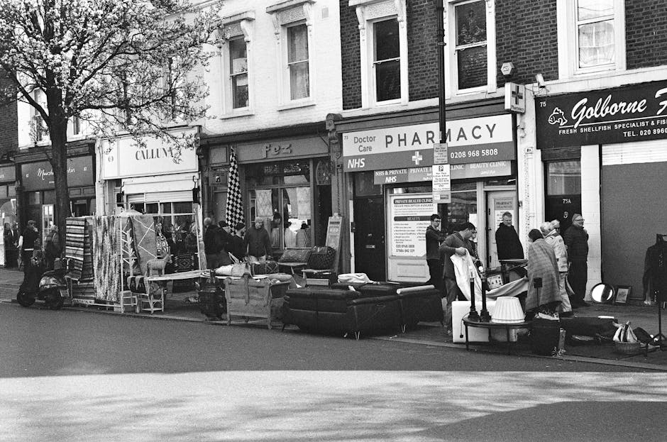 A busy street scene in Pimlico featuring various shops, including a pharmacy and seafood specialist, with several pedestrians, some wearing coats and hats, walking and standing on the sidewalk. Out fr